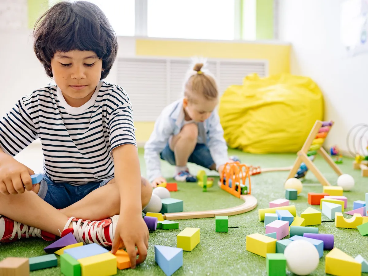 A young boy building with wooden blocks and a train set on a classroom rug